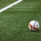 Ein Fußball mit rot-gelbem Muster liegt auf einem grünen Kunstrasenplatz nahe einer weißen Begrenzungslinie, bereit für das nächste Spiel des SV-Waldwimmersbach.