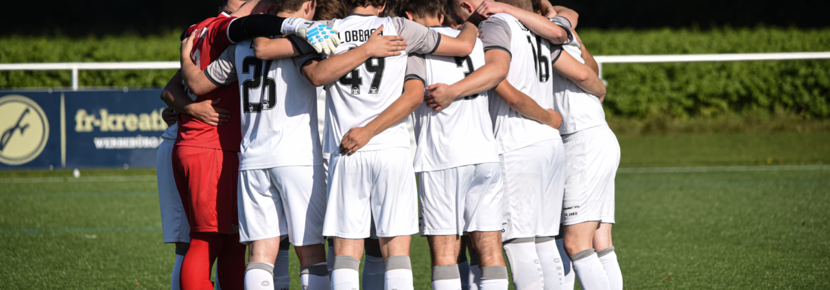 Eine Fußballmannschaft in weißen Trikots, vermutlich der SV Waldwimmersbach, steht dicht gedrängt auf einem grünen Feld, die Arme umeinander gelegt, bereitet sich auf ein Spiel vor oder bespricht Strategien, während das Sonnenlicht die Szenerie erhellt.