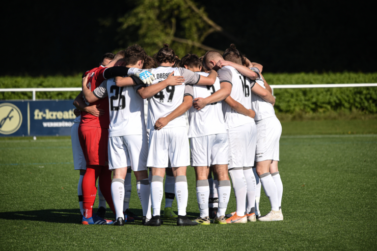 Eine Fußballmannschaft in weißen Trikots, vermutlich der SV Waldwimmersbach, steht dicht gedrängt auf einem grünen Feld, die Arme umeinander gelegt, bereitet sich auf ein Spiel vor oder bespricht Strategien, während das Sonnenlicht die Szenerie erhellt.