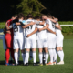 Eine Fußballmannschaft in weißen Trikots, vermutlich der SV Waldwimmersbach, steht dicht gedrängt auf einem grünen Feld, die Arme umeinander gelegt, bereitet sich auf ein Spiel vor oder bespricht Strategien, während das Sonnenlicht die Szenerie erhellt.